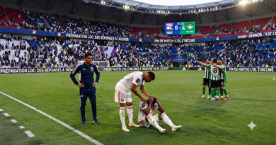 مدرب أولمبيك ليون ومدير فريقه (يسار) يشاهدان بلا مبالاة لاعباً من فريق ليون (يمين) جالساً على العشب، ممسكاً برأسه بيده، بينما يحتفل لاعبو فريق ريال بيتيس (في الخلفية) بهدفهم.