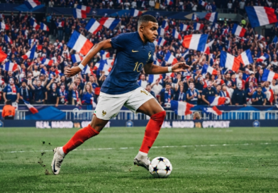 كيليان مبابي بقميص المنتخب الفرنسي يركض بالكرة على أرضية الملعب أمام جمهور يرفع الأعلام الفرنسية.