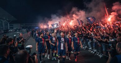 A wide, cinematic sports editorial photograph of Paris Saint-Germain players returning to Paris late at night after defeating Marseille, greeted by a euphoric crowd of supporters with red flares and smoke.