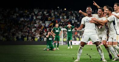 A dramatic, cinematic sports photograph capturing Real Madrid players celebrating a late winning goal against Elche in a packed stadium at night.