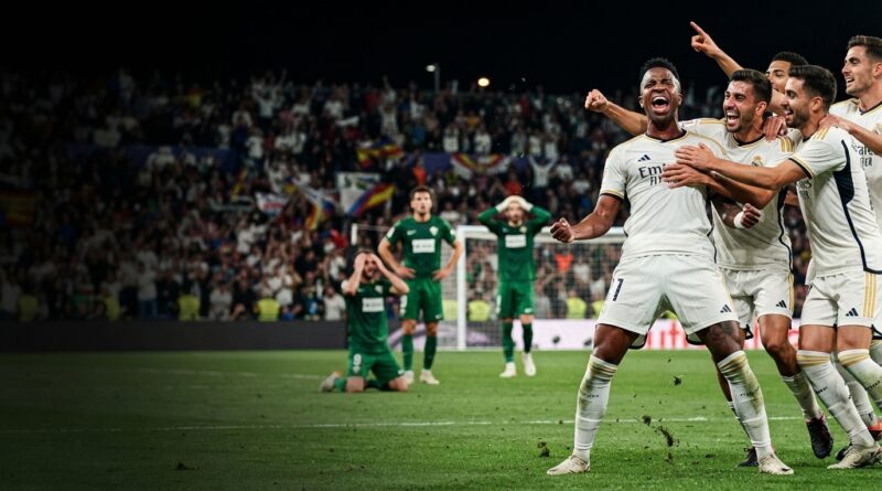A dramatic, cinematic sports photograph capturing Real Madrid players celebrating a late winning goal against Elche in a packed stadium at night.
