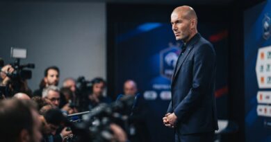 A cinematic sports editorial photograph captures Zinedine Zidane, in a dark navy suit with a subtle blue accent, standing composed and serious in a press conference room. Blurred foreground elements like microphones and cameras suggest intense media attention. The background is a sophisticated, unbranded French Football Federation-style backdrop with restrained lighting. The composition is 16:9, optimized as a hero image with Zidane to the right and negative space on the left.