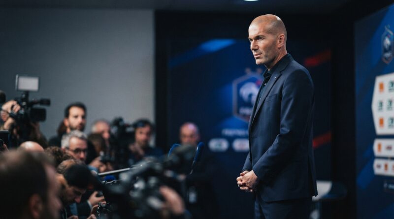 A cinematic sports editorial photograph captures Zinedine Zidane, in a dark navy suit with a subtle blue accent, standing composed and serious in a press conference room. Blurred foreground elements like microphones and cameras suggest intense media attention. The background is a sophisticated, unbranded French Football Federation-style backdrop with restrained lighting. The composition is 16:9, optimized as a hero image with Zidane to the right and negative space on the left.